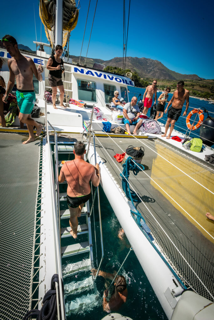 Un homme remontant à bord du catamaran par l'escalier central situé à l'avant entre les deux filets de détente, après une baignade dans la baie de Paulilles lors d'une Sortie bateau port-vendres