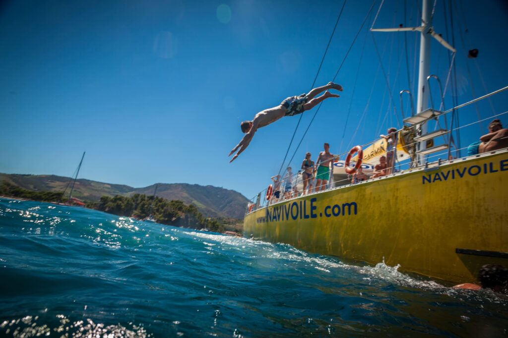 Photographie d'un homme effectuant un plongeon tête la première depuis le plongeoir du catamaran Navivoile dans les eaux turquoise de Paulilles lors d'une Sortie bateau port-vendres