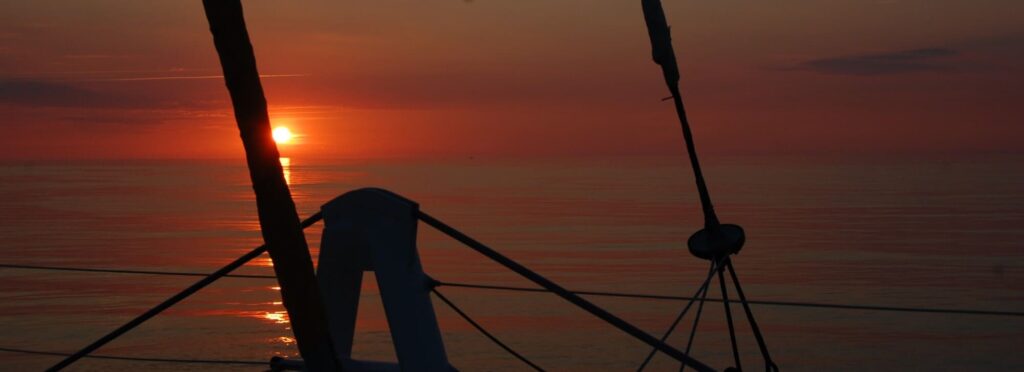 Photo d'une Sortie en Mer au Lever du Soleil à l'avant du navire. Le soleil se lève entre les étraves du catamaran Navivoile, à proximité des éoliennes flottantes du Golfe du Lion, lors d'une session d'observation de dauphin