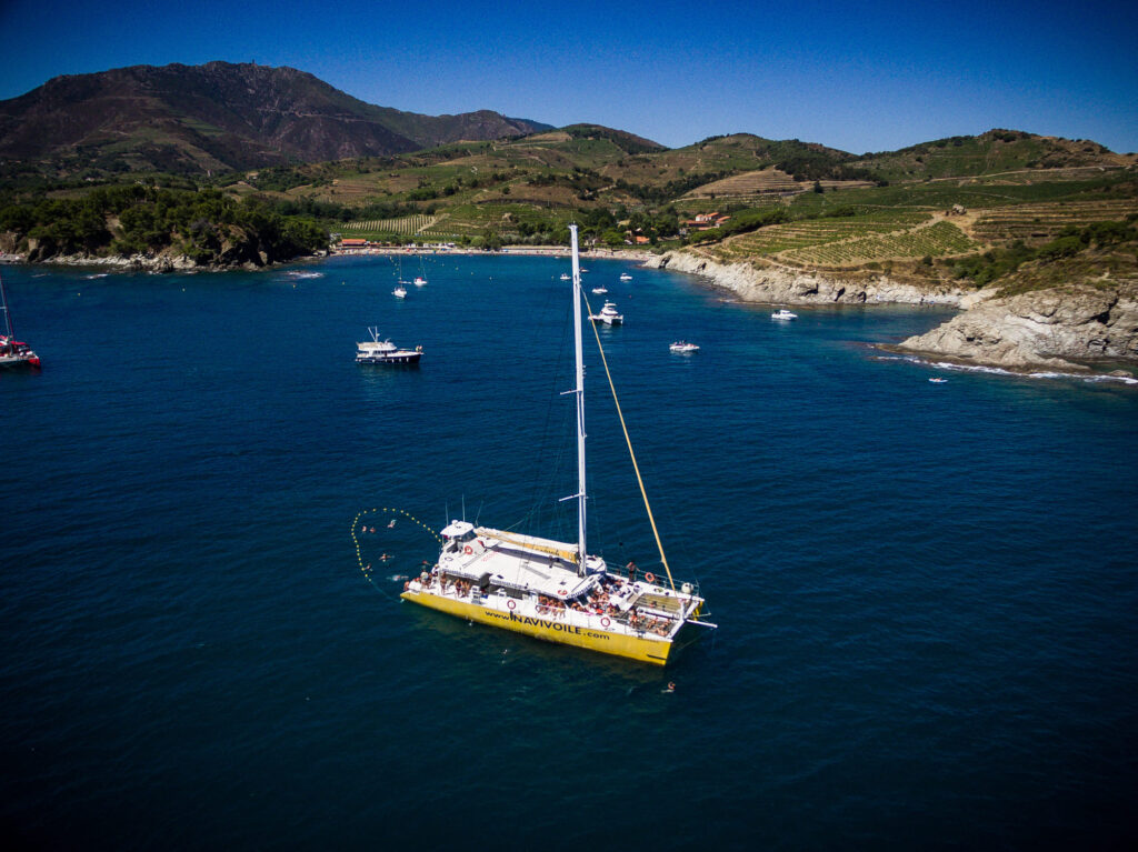 Vue aérienne par drone du catamaran Navivoile au mouillage dans les eaux turquoise de la baie de Paulilles, avec le massif des Albères en arrière-plan lors d'une Sortie bateau Canet