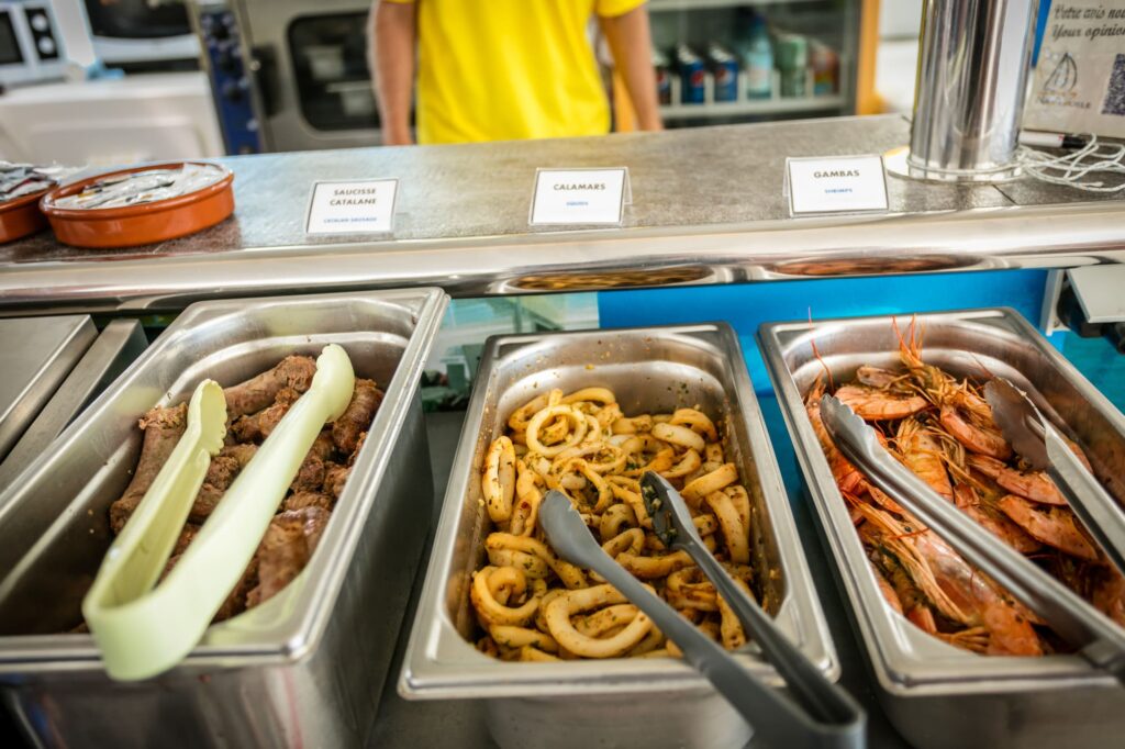 Plats de saucisses catalanes, gambas dorées et calamars fraîchement grillés sur le buffet du catamaran Navivoile au mouillage, lors d'une excursion maritime avec repas au départ de Canet