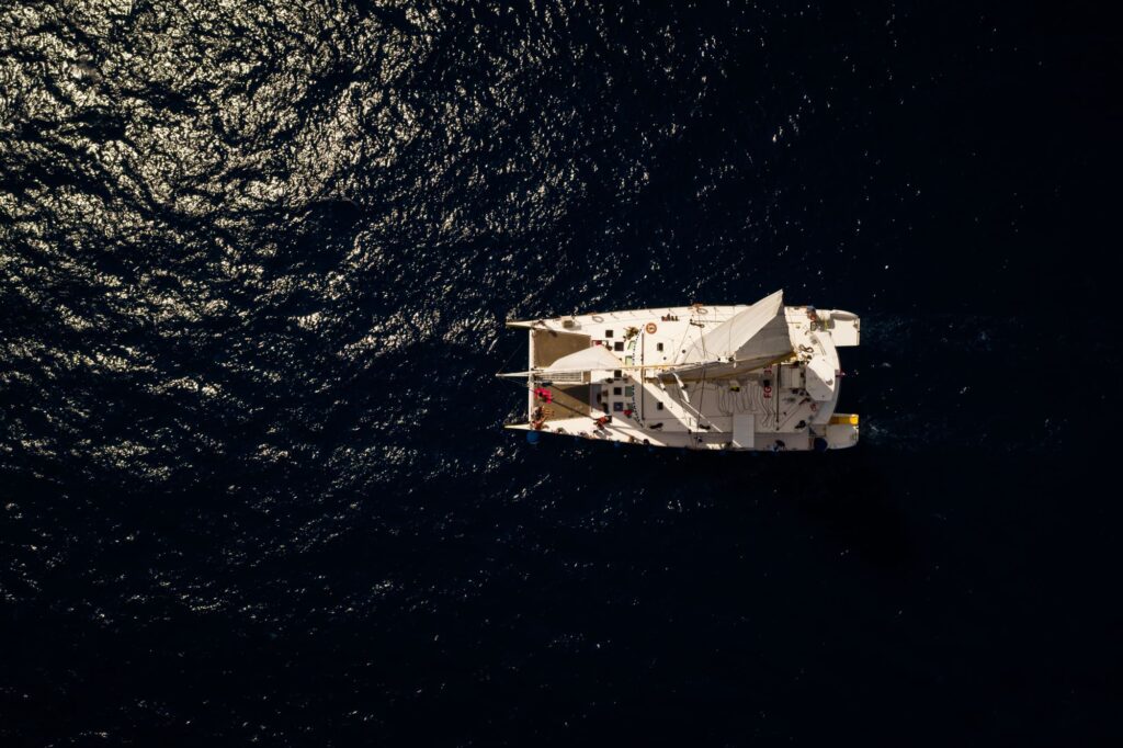 Photo du catamaran Navivoile prise au zénith par drone, montrant la grand-voile Oniria déployée sur une mer aux reflets argentés