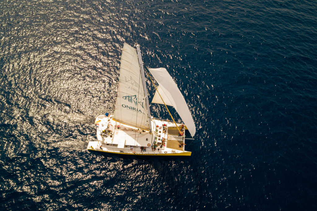 Vue de dessus par drone du catamaran Navivoile toutes voiles dehors avec le logo Oniria, glissant sur une mer scintillante à Canet