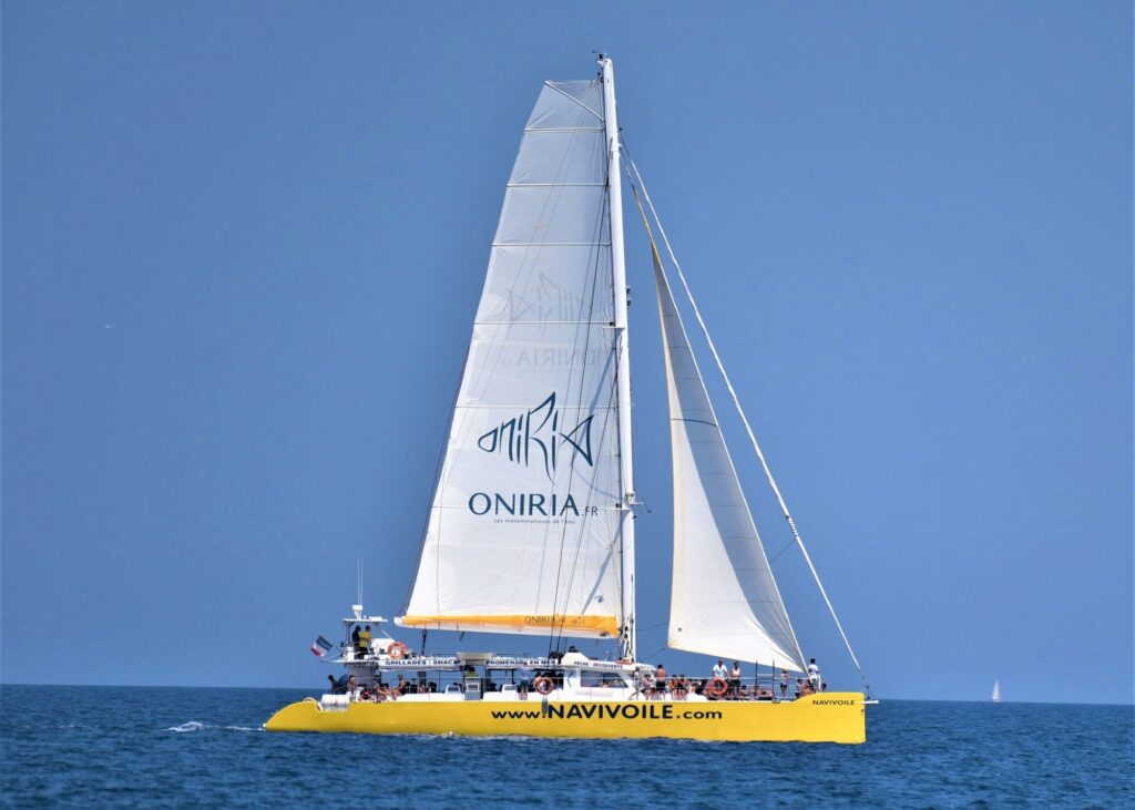 Le catamaran Navivoile naviguant sous voiles avec le logo de l'aquarium Oniria visible sur la grand-voile, passant devant le littoral de la station lors d'une Sortie en mer à Canet-en-Roussillon