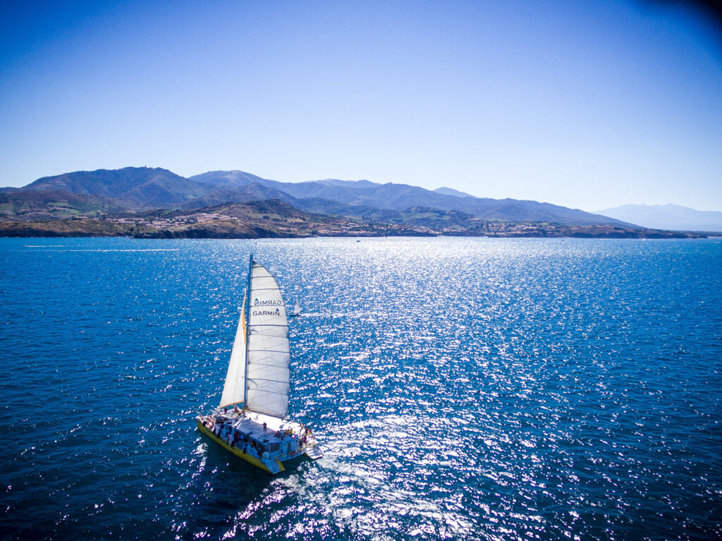 Le catamaran Navivoile naviguant sous voiles par légère brise vers Port-Vendres, avec la silhouette de Collioure en arrière-plan, lors d'une sortie bateau à la journée à Canet
