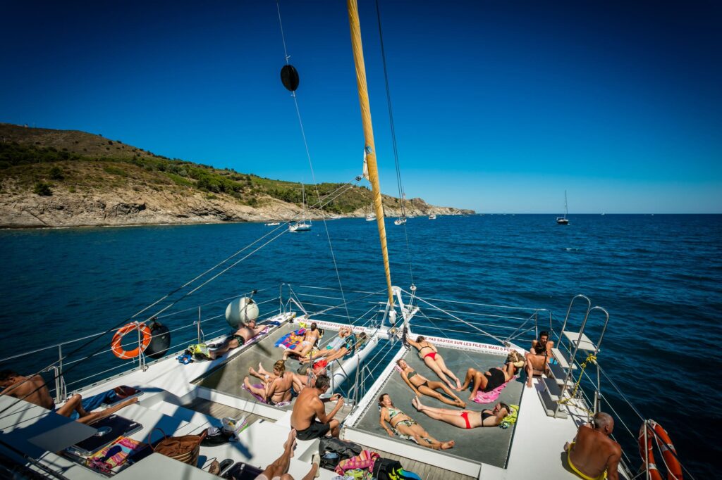 Passagers relaxés prenant le soleil sur les filets avant du catamaran Navivoile au mouillage à Paulilles, après un déjeuner grillades lors d'une sortie bateau à la journée à Canet