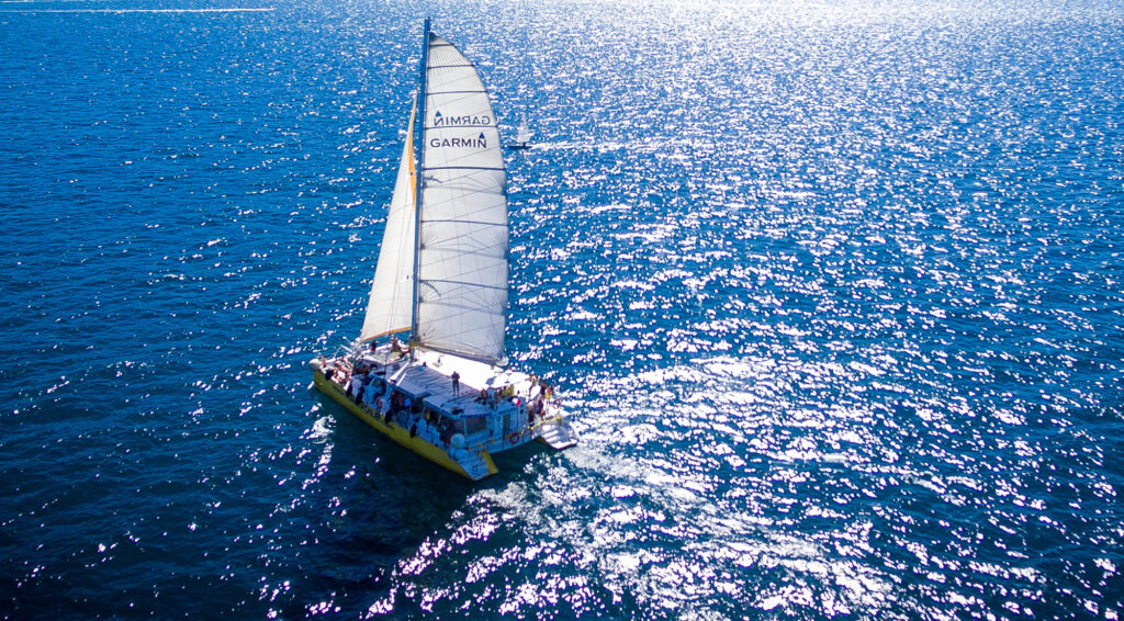 Vue aérienne à contre-jour du Navivoile toutes voiles dehors sur l'eau miroitante face au grau de l'étang de Canet-Saint-Nazaire