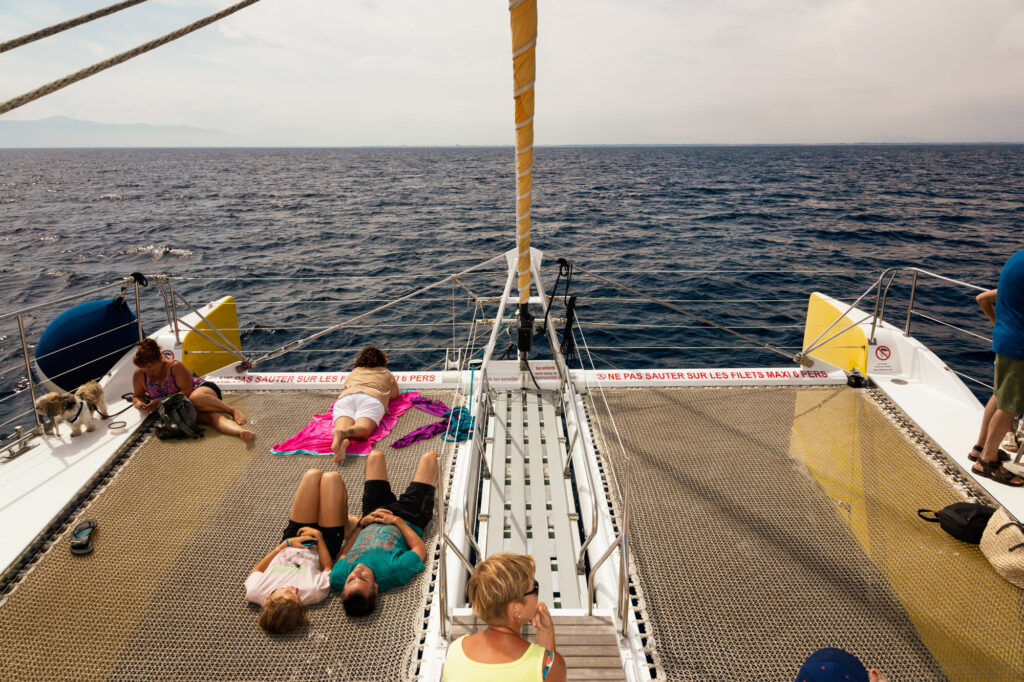 Vue vers l'avant du catamaran au pied du mât, montrant des passagers très reposés et zen profitant du calme, avec en arrière-plan la côte découpée et la colline du Cap Béar, lors d'une observation dauphins Port-Vendres avec le catamaran Navivoile