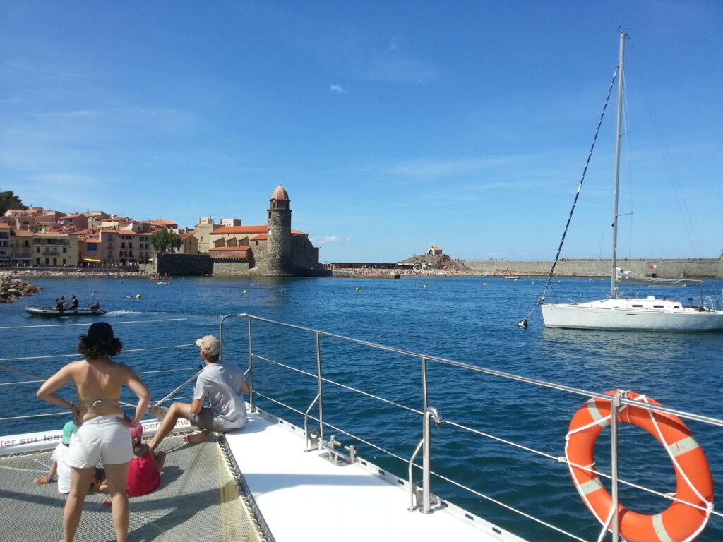 Passagers du catamaran Navivoile relaxés sur les filets avant, avec l'église de Collioure et son clocher iconique en arrière-plan