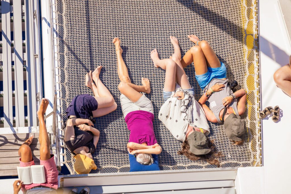 Vue plongeante d'une famille se relaxant dans les filets avant lors d'une Promenade en mer à Canet en Roussillon. Le catamaran Navivoile navigue sans moteur au-dessus des eaux claires face à la plage de Torreilles
