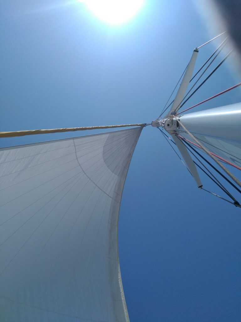 Vue en contre-plongée depuis le pied du mât lors d'une Promenade en mer à Canet en Roussillon. Le soleil brille à travers le gréement et les voiles du catamaran Navivoile en navigation devant le phare de Canet