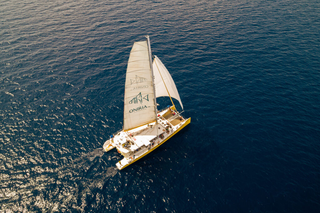 Vue aérienne par drone d'une Promenade en mer à Canet en Roussillon. Le catamaran Navivoile navigue sous voiles en contre-jour, créant des reflets argentés sur la Méditerranée au large de Saint-Cyprien