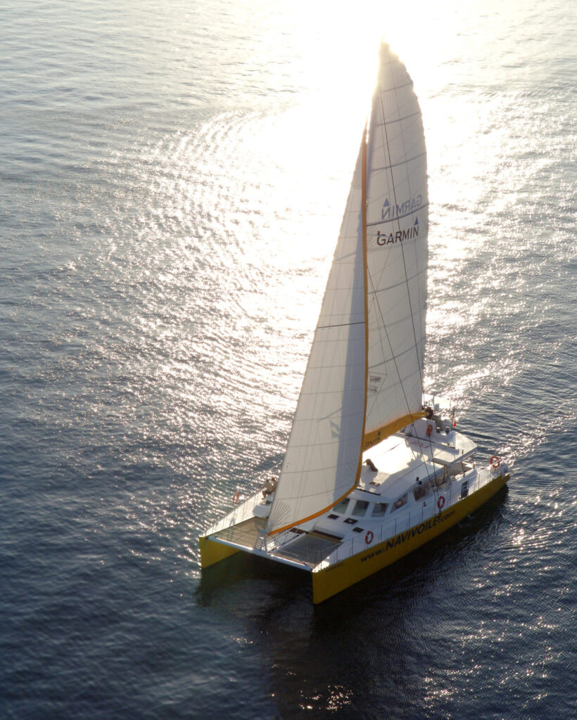 Photo en contre-jour matinal d'une Promenade en mer à Port-Vendres. Le catamaran Navivoile navigue sous voiles devant les falaises et les criques de Porteils, baigné par la lumière du soleil levant