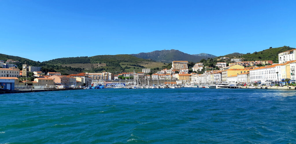Photo du catamaran Navivoile à quai dans le port de Port-Vendres. En arrière-plan, la place Castellane, la tour de l'Horloge à gauche et la tour de la Madeloc dominant les sommets des Albères