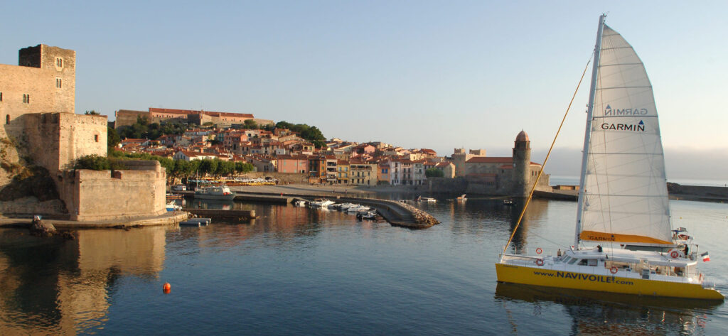 Photo d'une Promenade en mer à Port-Vendres. Le catamaran Navivoile navigue au plus près du clocher de l'église Notre-Dame-des-Anges et des remparts du Château Royal dans la baie de Collioure par calme plat