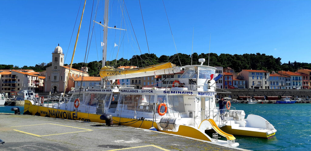 Photo du catamaran Navivoile amarré au quai de Port-Vendres avant l'embarquement pour une Promenade en mer à Port-Vendres. En arrière-plan, l'église Notre-Dame-de-Bonne-Nouvelle et son dôme bleu
