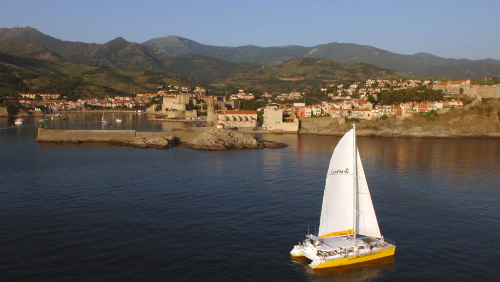 Photo d'une Promenade en mer à Port-Vendres. Le catamaran Navivoile navigue sous voiles en face de Collioure, avec la tour de la Madeloc dominant le massif des Albères en arrière-plan