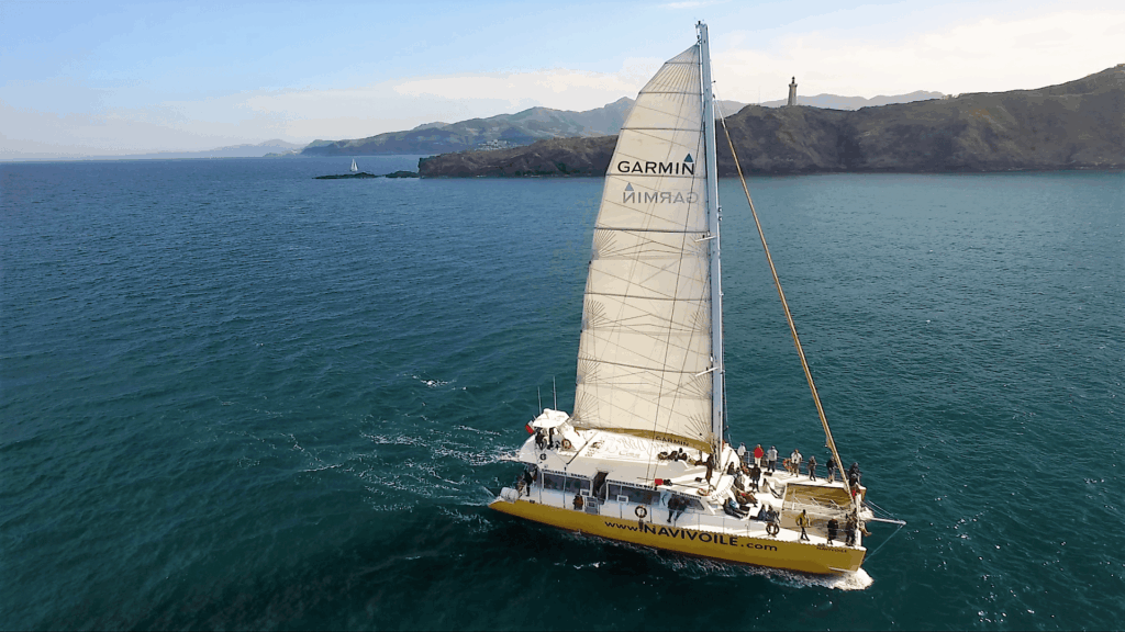 Vue aérienne du profil tribord du catamaran Navivoile naviguant sous grand-voile en avril, avec le phare et les falaises du Cap Béar en arrière-plan lors d'une Sortie en mer à Canet-en-Roussillon