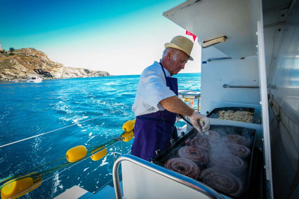 Bernard, père fondateur du Navivoile, prépare des gambas, saucisses catalanes et calamars sur les planchas. Scène de cuisine à bord lors d'une croisière grillades à Paulilles en face de la plage Bernardi avec le principal catamaran à voile à Canet-en-Roussillon et Port-Vendres