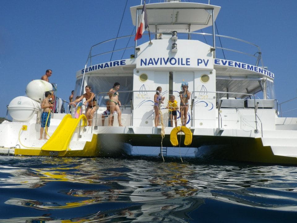 Un enfant nageant à l'arrière du catamaran Navivoile et utilisant le toboggan lors d'un ancrage dans la Baie de Paulilles. Scène de loisirs nautiques sur la Côte Vermeille avec le principal catamaran à voile à Canet-en-Roussillon et Port-Vendres