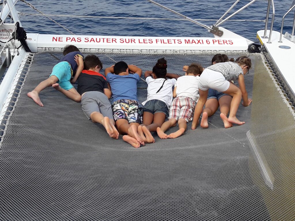 Des enfants de primaire regardent l'eau au travers des filets sécurisés à l'avant du Navivoile en navigation sous voiles devant la plage de Torreilles