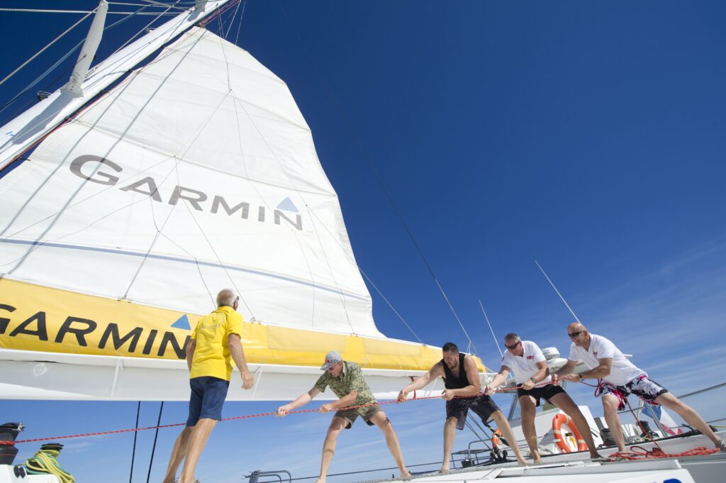 Effort collectif en team building d'une équipe qui hisse la grand-voile logotée GARMIN devant le port de Saint-Cyprien sur le NAVIVOILE